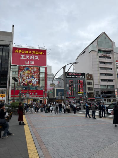 This media file captures a busy street scene in Taito, Japan, under an overcast sky during daytime. The image is dominated by multi-story commercial buildings adorned with numerous signs and advertisements, characteristic of a Japanese urban center.

A prominent building features the name "NAGAFUJI" on its facade and a distinctive, modern glass peak. Below it, another building houses a "LAWSON" convenience store and advertises "BILLIARDS DARTS." Further along, a large grey building also displays "NAGAFUJI."

A massive red billboard on the left side of the frame advertises "パチンコ&スロット エスパス 日扼" (Pachinko & Slot Espace Nitaku/Hishaku), featuring colorful anime-style characters, one making a large "V" sign. Another sign below it confirms "PACHINKO & SLOT ESPACE." In the mid-ground, a dark sign belongs to "ヨドバシカメラ マルチメディア上野2号店" (Yodobashi Camera Multimedia Ueno 2nd Store), listing departments such as "おもゃ・ホビー・玩具" (Toys, Hobbies, Games) and "ゲーコ・ゲームソフト" (Games, Game Software). A sign for "みばえ" is also visible.

The wide, tiled sidewalk is populated by numerous pedestrians walking, standing, or waiting. Among them are two police officers in uniform. A person wearing a red plaid skirt stands near the yellow tactile paving, while another person is seated on a low wall or bench in the foreground. Several taxis are visible on the street, suggesting active traffic. The overall impression is one of an active commercial district.
