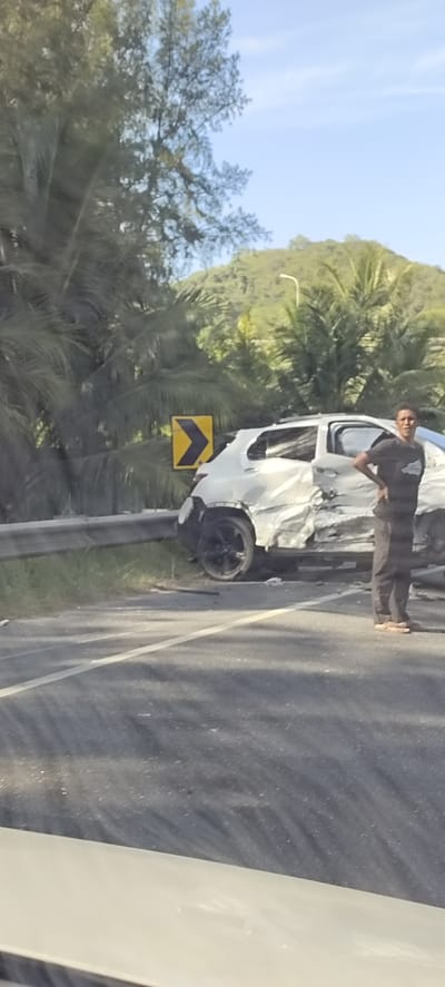 A white car with significant damage to its side is parked on the shoulder of a road, next to a metal guardrail. A person wearing a dark t-shirt and dark pants stands beside the car, with their hands on their hips, looking towards the camera. A yellow directional sign with a black arrow pointing to the left is visible behind the car. The scene appears to be in a rural or semi-rural area with lush green vegetation and palm trees on a hill in the background. The sky is clear and blue. The location is identified as Lamkruet, Indonesia.