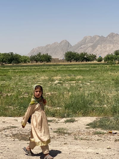 A young girl is walking on a dirt path in Deh-E Bagh, Afghanistan. She is wearing a light brown dress with gold trim, a patterned headscarf, and sandals. In the background is a grassy field, a line of trees, a wall, and mountains. The sky is clear.