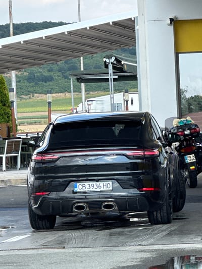 In Drazhevo, Bulgaria, a black Porsche Cayenne Turbo GT is parked in front of a building with a yellow facade and an awning. The vehicle's rear is visible, featuring a license plate that reads "CB 3936 HO". Next to the Porsche, a motorcycle with luggage strapped to the back is also parked, its license plate showing "PB A 3266". The ground is wet, indicating recent rain or cleaning. In the background, a row of tables and chairs suggests an outdoor seating area, possibly for a restaurant or cafe. Rolling hills covered in greenery form the backdrop. A security camera is mounted on the building.