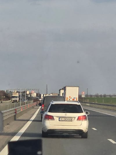 The image displays a multi-lane highway experiencing a traffic jam near Opalchenets, Bulgaria, under an overcast, light gray sky. The perspective is from inside a vehicle, looking forward, with a dark, out-of-focus object partially obscuring the bottom left of the frame. In the immediate foreground, a white Mercedes-Benz station wagon with a Bulgarian license plate "X 2434 KM" is stationary in the rightmost lane. Ahead of this vehicle is a dark-colored van, followed by a long queue of additional vehicles, primarily large trucks with white trailers, stretching into the distance.
On the left side of the highway, a gray metal guardrail with orange reflective strips runs alongside a concrete barrier. On the right, a similar gray metal guardrail separates the road from green and brown fields. Multiple circular regulatory road signs are visible in the distance on both sides of the highway, one clearly identifiable as a red circle with a white horizontal bar. Power line structures are faintly visible on the distant horizon. No individuals are explicitly visible.