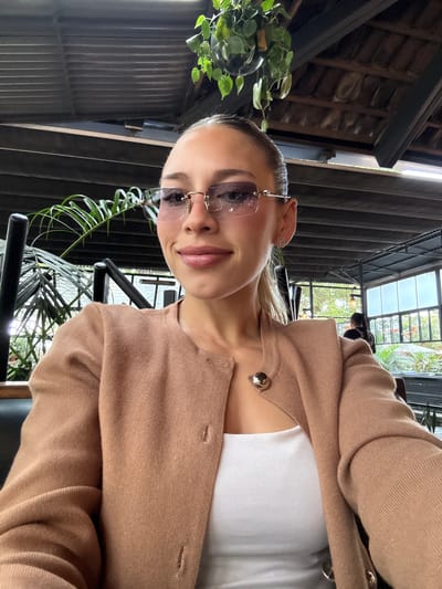 A woman in sunglasses and a tan sweater poses for a selfie indoors. She is wearing a white top underneath the sweater. The location appears to be a modern structure with dark metal beams and a partially visible ceiling. Green foliage hangs nearby. While the architectural style suggests a modern building, I lack enough data to confirm that the location is in the city of La Tijera, Mexico. No actions or interactions can be discerned from the photograph.