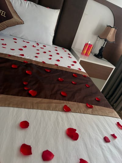 The image depicts a close-up, high-angle view of a king-sized bed prepared for a romantic occasion in Cam Lâm, Vietnam. The bed is covered in white linens, with red rose petals scattered across the surface, concentrated on a darker brown runner. A brown decorative pillow with "HOTEL" printed on it is positioned on the left side of the bed. To the right of the bed, a nightstand holds a decorative lamp with a beige shade and two cylindrical pink candles. The overall scene suggests a hotel room setting in Cam Lâm, Vietnam, prepared for a special event.