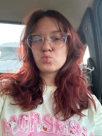 A young woman with long, wavy, dark red hair and glasses is in the driver's seat of a car, facing forward. She is making a puckering gesture with her lips and wearing a light-colored t-shirt with pink text and graphics. The background shows a window with a blurry view of what appears to be a street or another car, suggesting the vehicle is in motion. A car door handle and part of a seatbelt are visible on the right side of the frame. The scene is indoors, within a vehicle. There is no specific indication of the location within the city of Ленин, Kyrgyzstan.
