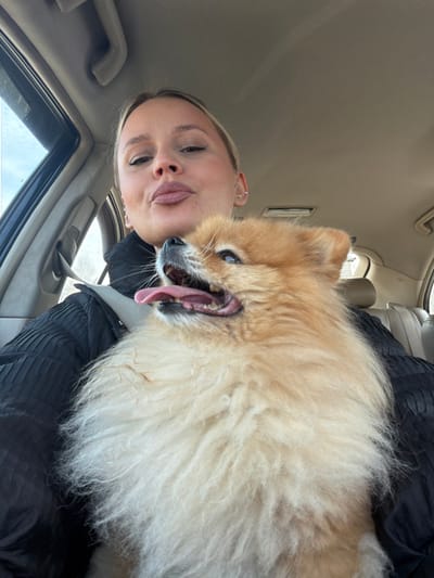 A female individual and a small, fluffy dog are depicted inside a vehicle in Makarovo, Russia. The female, positioned in the foreground, has light hair pulled back, visible ear piercings, and is looking towards the camera with a pursed-lip expression. She wears a dark, possibly black, textured garment, and a gray seatbelt is fastened across her chest. The dog, a Spitz-type breed with predominantly orange and cream-colored fur, is held on her lap or chest. Its mouth is open, tongue extended, and its eyes are directed upwards and to the right. The vehicle's interior features light-colored (beige/tan) upholstery on the ceiling and visible headrests. Through the vehicle's left-side window, a blurred outdoor scene with light, possibly overcast, sky and indistinct vegetation is visible.