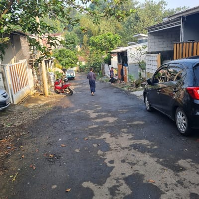 The image shows a narrow, paved street in the city of Kecamatan Tamansari, Indonesia, located at the coordinates -6.635 latitude and 106.738 longitude. The street appears to be wet, likely from recent rain, and has a few scattered leaves on its surface. There are three individuals walking along the street; one is wearing a long-sleeved, dark-colored shirt and long pants, walking away from the camera, while the other two, who are both wearing light-colored shirts and pants, are walking toward the camera. They are situated near the entrance of a small residential area. To the left of the street is a building with a wooden fence in the front, and there is a car parked in front of it. There is also a motorcycle with a red body and a black seat parked on the left side of the street, near the building. A gray car is parked on the left side of the street further down. On the right side of the street, a black car is parked. The houses on the right side of the street are white with a gray roof and have several windows, some of which have blinds or curtains.  Dense foliage surrounds the houses.  The sky is not visible in the image.