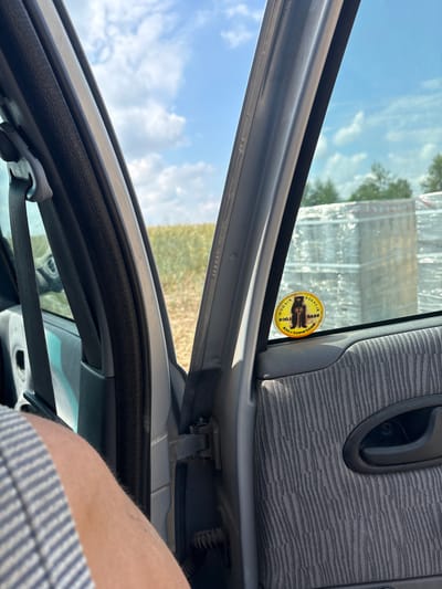 The image shows a view from inside a car, looking out through the open passenger door. A portion of a person's arm and shoulder, clad in a blue and white striped shirt, is visible in the lower left of the frame. The car's interior, including the door frame, seatbelt, and fabric-covered door panel with a black door handle, is also visible. On the exterior of the car window, a circular yellow sticker with a black bear illustration and text that appears to read "BEAR LOCK" is affixed. Outside the car, a field of crops, likely grain, stretches towards the horizon under a partly cloudy blue sky. In the middle ground, several large, palletized loads wrapped in clear plastic sheeting are visible, suggesting a commercial or agricultural context. The scene appears to be located in a rural or semi-rural area, possibly on the outskirts of Opatów, Poland, given the presence of agricultural fields and commercial goods.