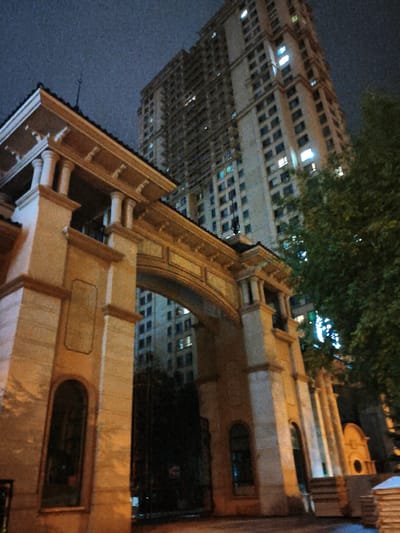 This is a nighttime, eye-level, outdoor, full-shot photograph of a large, modern, beige apartment building with many illuminated windows, situated behind a grand, arched entranceway. The entranceway is made of stone and features columns and decorative elements. To the right of the apartment building, a large, dark green tree is visible. The sky is dark and appears overcast. The image has a grainy texture, suggesting it was taken in low light conditions with a digital camera. The location is identified as the city of Jixi, China. There are no people or discernible actions captured in the image. The overall scene depicts a residential area at night, with the architecture of the apartment building and the entranceway being the prominent subjects.