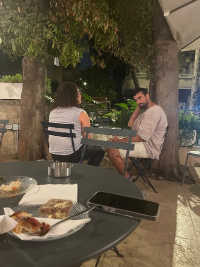 An outdoor night scene in West Jerusalem, Israel, depicts two individuals seated at separate dark round tables within a café or restaurant patio. A woman with dark, curly hair, wearing a light gray sleeveless top and dark pants, is seated with her back to the camera, facing a man. The man, with dark hair and a beard, wearing a light-colored loose-fitting short-sleeved top and light shorts, sits opposite her, facing towards the camera with his left hand positioned behind his head. Both are seated on dark metal chairs.

On the table between the two individuals are two wine glasses, each containing a small amount of white wine. In the foreground, a dark round table holds a silver-colored ashtray, a black smartphone in a clear case lying screen-down, and three small clear glass plates. These plates contain remnants of partially consumed food items, including a pastry with a crumbly topping, a savory pastry with red sauce, and another mostly consumed item. White paper napkins are positioned beneath some plates.

The environment is dimly lit by artificial light, set beneath large trees with dark foliage. A prominent tree trunk stands between the two seated individuals. The ground is paved with stone. Distant, dimly lit architectural details are visible in the background, and a white patio umbrella is partially visible in the upper right.