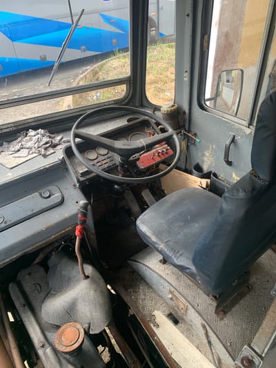 An interior view of an aged bus driver's cabin in Koggala, Sri Lanka, shows a worn black vinyl driver's seat with visible tears, positioned next to a metal floor with a diamond plate pattern. The dashboard features a dark steering wheel with the "TATA" logo, multiple gauges, and a red panel with several toggle switches and buttons. A gear shifter, wrapped with red and black tape, is located to the left of the steering column. A rag is placed on the dashboard to the left of the steering wheel. The windshield, showing a blurry view of another bus with blue and gray detailing and a grassy embankment, is equipped with a single windshield wiper. The cabin walls are gray and appear weathered. A black side-view mirror is visible on the right-hand side, reflecting a light-colored wall. The engine compartment is partially visible beneath the dashboard, with some metallic and gray components. The overall scene suggests a neglected and potentially non-operational vehicle.