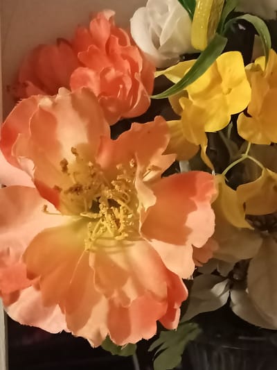 The image shows a close-up of artificial flowers, primarily in shades of peach and yellow, with hints of white and green foliage. The lighting is soft and slightly diffused, creating a warm tone across the petals. The arrangement appears to be part of a larger bouquet or display, with some flowers in sharp focus and others slightly blurred in the background. The overall impression is one of a still life, emphasizing the texture and color of the floral elements. There is no indication of the city of Amarillo, United States of America, nor any other specific location or environmental context within the image itself.