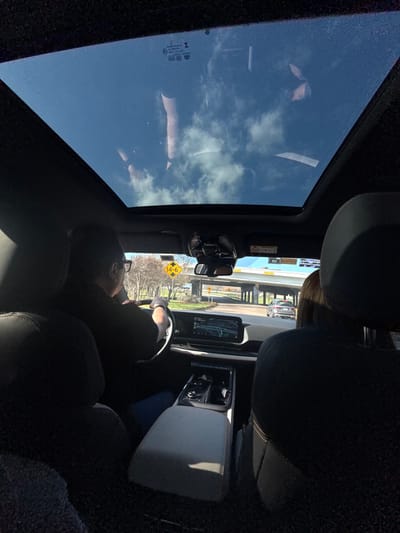 Inside a car, a driver in a black shirt holds the steering wheel, looking toward a roadside sign. The car travels under a bridge. Sunlight streams in through the sunroof, reflecting clouds. The vehicle's interior features black seats and a light gray center console. A navigation display is visible. Another passenger is present in the backseat.