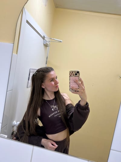 A woman is taking a selfie in a bathroom mirror. She is holding a pink and patterned iPhone. The bathroom has yellow walls and white tiles. A white metal toilet flusher arm is visible to the left of the frame. A toilet paper holder is partially visible in the bottom right of the frame. The woman is wearing a dark brown crop top and a matching zip-up jacket. The text on her shirt reads "HUDSON NICE." Her hair is long and brown, and she is wearing a hair clip on one side. The location is unspecified.