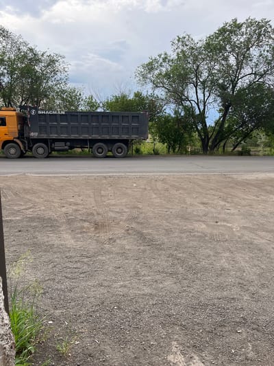 A horizontal, low-angle shot captures a large, dark gray dump truck parked on the side of a paved road. The truck, identified by the text "SHACMAN" and "1017" on its side, appears to be in a work-related setting. Its massive tires and utilitarian design suggest it is used for hauling materials. The truck is positioned next to a dirt or gravel area, which shows tire tracks, indicating recent vehicle activity.

To the left of the road and truck, a line of lush green trees with dense foliage creates a natural border. Beyond the trees, the sky is a mix of blue and white clouds, suggesting daytime with partly cloudy weather. The sunlight appears to be diffused, possibly indicating it's not the peak of midday.

The scene is located in Termirtaw city administration, Kazakhstan. The overall impression is one of an industrial or logistical operation, with the dump truck being the central object of focus. There are no people visible in the frame, and the environment appears functional rather than recreational. The presence of the trees and sky provides a natural contrast to the man-made machinery. The image is oriented such that the truck is viewed from the side, with its bed facing upwards towards the sky.