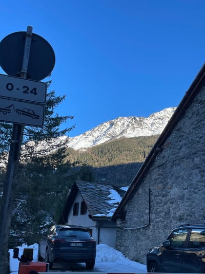 The image depicts a serene winter scene in the mountainous region of Courmayeur, Italy, under a clear, bright blue sky. A towering, snow-capped mountain range dominates the background, with evergreen forests covering its lower slopes, parts of which are in shadow.

In the mid-ground and foreground, traditional stone architecture is visible, including a prominent stone wall on the right side. Below this, a small house with light-colored walls, dark shutters, and a dark, textured roof covered in snow adds to the alpine village setting.

Several objects are present: two dark-colored SUVs are parked. One is partially visible near the upper stone wall, reflecting the sky. The other is parked near the small house, and its Italian license plate, "GT 967LX," is clearly visible. Patches of snow are scattered on the ground around the buildings and vehicles.

In the bottom left, a pole holds two white rectangular road signs. The left sign displays "0-24" vertically, while the right sign shows an icon of a car, possibly indicating parking information or road conditions.

The scene captures a peaceful moment on a sunny day, likely morning or early afternoon, given the crisp light and distinct shadows. No people are visible, emphasizing the natural beauty and quietude of the snowy landscape. The overall impression is one of tranquility in a picturesque mountain setting.