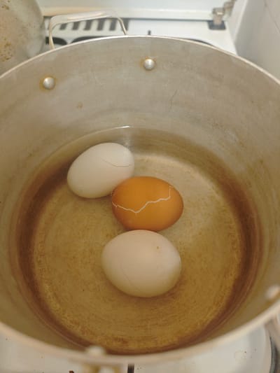Three eggs, two white and one brown, are submerged in water in a large, stained metal pot. The eggs are positioned in the center of the pot. The white egg on the left has a visible crack in its shell. The brown egg, situated slightly in front of the white one, also exhibits a crack. The third white egg is positioned at the bottom of the pot. The pot appears to be on a stovetop, with part of the white stovetop visible at the bottom of the frame. The background is out of focus, showing the upper portion of the stovetop and a metallic handle. The lighting suggests indoor illumination. The image was taken in Volkhov, Russia.