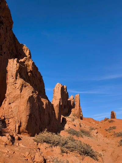This vibrant image captures a dramatic desert landscape dominated by towering red rock formations under a clear blue sky, characteristic of the Tosor region in Kyrgyzstan, particularly "Skazka Canyon" (Fairytale Canyon). On the left, a massive, rugged red cliff face rises sharply, showcasing intricate erosion patterns and deep shadows that highlight its textured surface. Extending into the middle ground are several slender, spire-like rock pillars, sculpted by natural forces, their peaks jagged and weathered. These formations cast long, dark shadows across the reddish-brown terrain, indicating strong sunlight.
The ground is composed of rust-colored earth and small rocks, typical of an arid environment. Sparse, dry bushes and low-lying shrubs with muted green and brown foliage are scattered across the foreground and mid-ground, providing a touch of vegetation against the predominantly red landscape. In the distance, on the right, further smaller rock formations suggest the expansive nature of the area.
The sky is a brilliant, unblemished blue, with only the faintest wisps of white clouds visible in the upper right, denoting clear and sunny weather. The strong contrasts between light and shadow on the rock faces suggest the photograph was taken during midday or early afternoon, under direct sunlight. There are no people, animals, or any specific activities visible within the frame, portraying a serene and untouched natural scene. No text is present in the image.