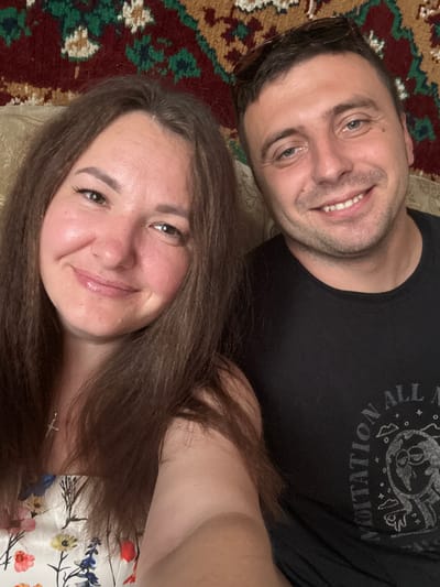 The image is a selfie of a man and a woman. The man is on the right, smiling and wearing a black t-shirt with a graphic design. The woman is on the left, also smiling and wearing a white dress with a floral pattern. They are both close to the camera, and their faces fill most of the frame. Behind them, a patterned rug with red, green, and yellow colors is visible. The setting appears to be indoors, possibly a living room. The man has brown hair and blue eyes, while the woman has long brown hair and brown eyes. The image quality is good, with natural lighting. There is no indication of the specific location in Хаціслаўскі сельскі Савет, Belarus, beyond the general appearance of a domestic setting.