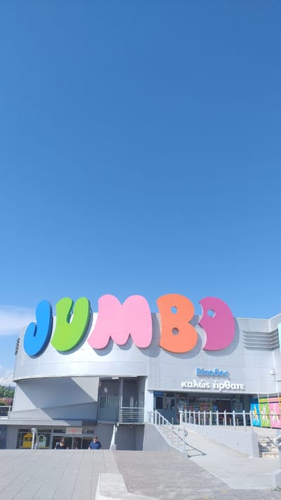 A vertical, eye-level, exterior shot depicts the facade of a Jumbo store under a clear blue sky in Pylaia Municipal Unit, Greece. The prominent, colorful "JUMBO" logo, with letters in blue, green, pink, orange, and purple, spans the upper portion of the building. Below the logo, a gray, modern building with multiple entrances and a ramp leading up to one of the entrances is visible. Two people are standing near the lower entrances, one near a yellow doorway on the left, and another in a blue shirt in the center. The ground in front of the building is paved with gray concrete, with a concrete barrier in the foreground. The overall impression is a bright, sunny day at a large retail establishment.