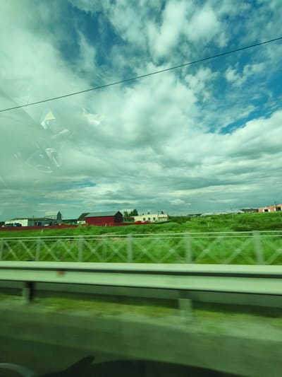 The image captures a view from a vehicle, likely a car, looking out through a tinted window. The sky is a prominent feature, filled with dramatic, billowing clouds and patches of bright blue. A thin, dark wire is visible stretching horizontally across the upper portion of the frame, possibly a power line or telephone wire.

In the middle ground, there is a rural landscape with several buildings. A large, red barn-like structure stands out, with a darker roof. To its left, a smaller, light-colored building with a grey roof is visible, and further left, a white building with some industrial-looking structures. The buildings are situated amidst lush green fields and some trees. A metal fence with a diamond pattern separates the field from a road or pathway in the foreground.

The foreground is dominated by the interior of the vehicle, reflected on the window. A steering wheel and part of the dashboard are visible, indicating the perspective of the photographer is from the driver's seat. The lower part of the image shows a blurred reflection of the road and possibly the shadow of the vehicle's occupants.

The presence of rural buildings, the style of the fence, and the overall landscape suggest a location in a rural area. While the prompt mentions Issad, Russia, there are no specific visual cues within the image that definitively identify this precise location. The scene depicts a typical roadside view from a moving vehicle in what appears to be a rural or semi-rural setting during the daytime.