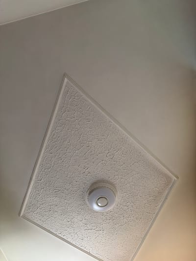 The image shows a ceiling in Umarevtsi, Bulgaria. A decorative square panel with a heavily textured surface is attached to the ceiling. A round, white light fixture is mounted in the center of the panel. The ceiling itself appears to be plain and painted a light color. The panel is oriented at an angle to the camera.