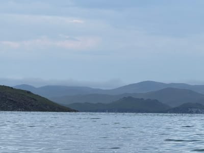 The image depicts a serene, overcast landscape dominated by a body of water in the foreground and a series of rolling, mist-shrouded hills in the background. The water exhibits subtle ripples, reflecting the muted tones of the sky. The hills, layered in varying shades of blue and green, create a sense of depth and distance. Vegetation appears sparse, with some grassy slopes visible on the closer hills. The sky is a soft, cloudy blue, suggesting a cool or temperate climate. The scene evokes a sense of tranquility and natural beauty, characteristic of a remote, undeveloped area. The location is identified as Sarma, Russia, implying a setting in a region known for its natural landscapes.