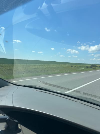 The image captures a view from inside a vehicle, looking out through the windshield. The windshield itself has some visible smudges and a crack running through the lower central portion. Outside, the scene depicts a sunny day with a bright blue sky dotted with fluffy white clouds.

Dominating the mid-ground is a vast, flat expanse of green fields, likely agricultural land, stretching towards the horizon. A paved road, marked with a white line, runs diagonally from the lower right corner towards the center, indicating the vehicle is moving along a highway. The road appears to be in good condition. Tire tracks are faintly visible on the road surface. In the far distance, faint outlines of power lines or poles can be seen against the horizon.

The foreground is occupied by the interior of the car, specifically the dashboard. The dashboard is a dull gray plastic with a textured surface along the top edge. Part of a side mirror assembly is visible on the left side of the image, mounted near the A-pillar.

Without specific landmarks within the image, it is impossible to pinpoint the exact location within the city of Қазақ, Kazakhstan. However, the landscape suggests a rural or semi-rural setting characterized by agricultural fields and open spaces, typical of many areas in Kazakhstan. The overall impression is one of travel through a scenic, open landscape on a clear day.