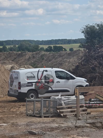 The image shows a white van parked on a dirt construction site, likely in Piecnik, Poland, given the location context. The van has branding for "RentWil" with a phone number "600 97 57 27" and the slogan "Podnosimy dużo i wysoko" (We lift a lot and high). The van is decorated with graphics of construction equipment, specifically a loader. A metal cage with some contents is placed next to the van, and wooden planks are part of the construction setup. The background reveals a hilly landscape with trees and fields under a partly cloudy sky. The lighting suggests it is daytime, possibly late morning or afternoon. The overall scene depicts a typical construction or equipment rental scenario. The license plate of the van is partially visible and reads "ELM K34".