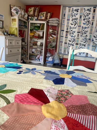 A close-up shot focuses on a partially completed quilt laid out on a table. The quilt features a large, stylized flower design with fabric petals in various colors and patterns, including pink gingham, floral prints, solid red, polka dots, and stripes. Another, smaller blue flower motif is visible in the background on the same quilt. The background of the image reveals a room with shelving units filled with fabric and craft supplies, and a large framed quilt hanging on the wall. A white chair with a teal measuring tape draped over it is positioned near the table. The setting appears to be a craft room or sewing studio.