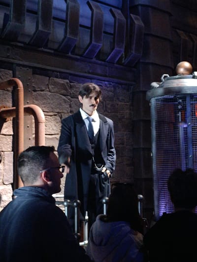 The image shows a dimly lit interior space with an industrial aesthetic. A man dressed in a dark suit, white shirt, and tie, with a thin mustache, stands in the center, his left hand in his pocket, and a pocket watch chain visible. He appears to be giving a presentation or performance to a group of spectators. The audience, consisting of at least three individuals, is positioned in front of him, their backs to the viewer. To the left of the presenter, there's a rusty, curving pipe structure. To his right is a cylindrical cage-like structure, with a metallic sphere on top. The background consists of stone-like walls and dark, geometric shapes above, giving the impression of a factory or laboratory setting. There is no indication this location is in Rust, Germany.