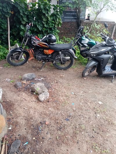 The scene is located in Pameungpeuk, Indonesia at latitude -7.668 and longitude 107.694. It is a dirt lot, with a black Yamaha motorcycle parked on the left. The motorcycle has a black helmet on the seat. On the right is a green and black moped parked on the dirt. Both vehicles are facing toward the right. The dirt lot has sparse vegetation growing on it, including grass and small shrubs. There are a few loose rocks and debris on the ground. There are no people visible in the scene.  There is a brick wall in the background with some writing on it. The scene is dimly lit.
