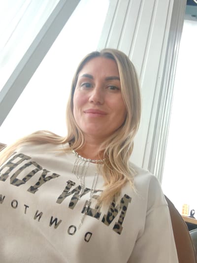 The media file shows a medium shot of a woman with blonde hair. She is looking at the camera with a slight smile. She is wearing a light-colored sweatshirt with a leopard print pattern on the letters that spell out "BOY M". Below this, another line of text appears to read "NOTHWO D". She is also wearing a delicate silver necklace with pearls.
The setting appears to be indoors, with natural light coming from large windows or glass doors. The background is bright and somewhat blown out due to the light. Vertical white elements, possibly part of the building's architecture or blinds, are visible on the right side of the image, and a diagonal white element is on the left. A hint of a brown chair is visible in the lower right corner. The overall mood is casual and relaxed. There are no clear indicators of the time of day or weather, but the bright light suggests daytime. The location is identified as Amursk, Russia.