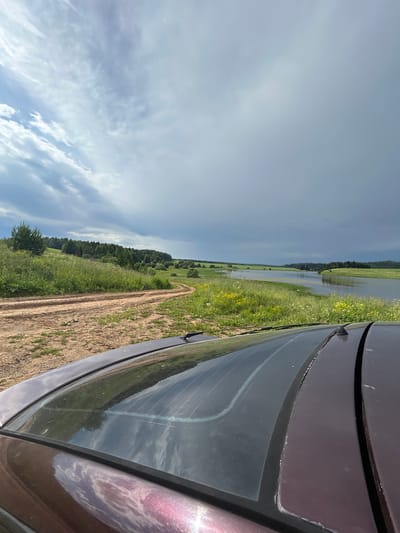 The image captures a rural landscape near Verkh-Rozhdestvo, Russia. In the foreground, the dark purple roof and rear window of a car are visible, angled towards the right. The rear window reflects the sky and surrounding greenery. A dirt road curves through a grassy field dotted with yellow wildflowers, leading towards a body of water. The water, likely a river or lake, stretches into the distance, bordered by green fields and a dense line of trees on the horizon. The sky is dramatic, with a mix of bright clouds and darker, stormier formations overhead. The overall scene conveys a sense of natural beauty and a journey through the Russian countryside.