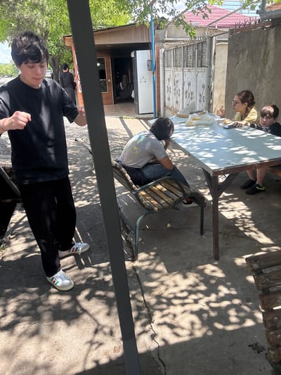 A medium shot shows four people gathered around a white picnic table outside a building in Badiauri, Georgia. The building appears to be a small shop with a red-tiled roof and a metal gate. In the foreground, a young person in a black t-shirt and black pants stands, facing the camera with fists clenched. To the right of the table, two children are seated, a girl with glasses eating and a boy in a black t-shirt looking at the camera. A third person, wearing a grey t-shirt and jeans, is seated on a wooden chair, facing away from the camera. The table has food and drinks on it, and the ground is paved. The scene is illuminated by sunlight filtering through the trees, casting dappled shadows.