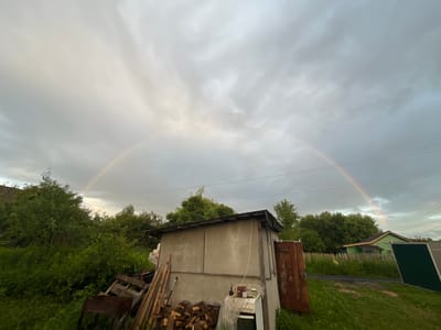 The image captures a cloudy sky with a faint double rainbow stretching across it, located in the village of Klobushnevo, Russia. In the foreground, a dilapidated concrete shed stands to the right, with a rusty metal door and various discarded items around it, including stacked firewood, wooden planks, and a rusty barbecue grill. Lush green foliage and trees surround the shed and extend into the background, where a small green house with a dark roof is visible. Power lines are strung across the sky above the trees. The overall scene suggests a rural, perhaps slightly neglected, outdoor setting after a rain shower.