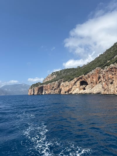 The image shows a coastal scene with a tall, rocky cliff face covered in vegetation on the left side. The cliff has a cave opening near the center. The right side of the image is dominated by dark blue water, showing some waves and white foam. The sky is visible in the upper left corner, with blue and white clouds. The location is Kumluca, Turkey.