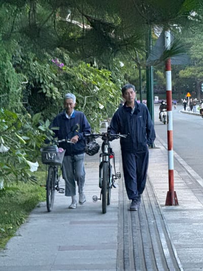 Two elderly men are walking on a sidewalk in Xuan Huong Lake, Da Lat, Vietnam. The man on the left has grey hair and is wearing a blue jacket and grey pants. He is pushing a bicycle with a basket in front. The man on the right has dark hair and is wearing a blue jacket and blue pants. He is walking next to his bicycle. The sidewalk is made of concrete with textured strips. There are trees and greenery on the left side of the sidewalk, and a road with traffic on the right. A red and white striped pole is standing on the sidewalk. In the background, there are other people walking and riding motorcycles. The scene appears to be during the day, with overcast lighting.