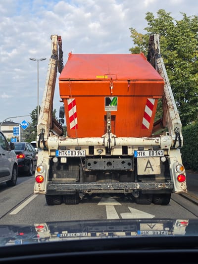 A large, orange dumpster truck is stopped in traffic on a street in Rüsselsheim am Main, Germany. The truck has a white lifting mechanism attached to its front and a license plate that reads "MTK DD 141". A smaller license plate below it shows a large "A". The truck has red and white reflective stripes on the back. To the left of the truck, a white car is stopped in traffic. In the background, trees and buildings are visible, along with a blue ARAL gas station sign with fuel prices. The sky is partly cloudy. The image is taken from the perspective of another vehicle in traffic.