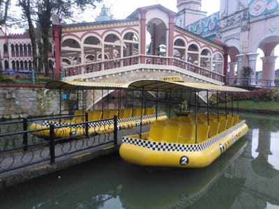 The scene, located in Ciudad de San José, Costa Rica, depicts a canal setting with two unoccupied, yellow watercraft designed to resemble taxis. In the foreground, these inflatable-style boats are moored or floating near a concrete dock featuring a black metal railing. Each boat is equipped with multiple yellow seats, a black canopy with a "TAXI" sign, and a black and white checkered pattern along its sides; the nearest boat bears the numeral "2" on its bow.
In the midground, an ornate, multi-arched bridge with a beige base and red structural elements spans the water, exhibiting a decorative railing. The background reveals elaborate, multi-story buildings characterized by intricate architectural details, including numerous arched windows and decorative facades in red, beige, and light blue tones. Green foliage and trees are visible on the banks and in the distant background, under an overcast sky. No individuals are present in the frame.