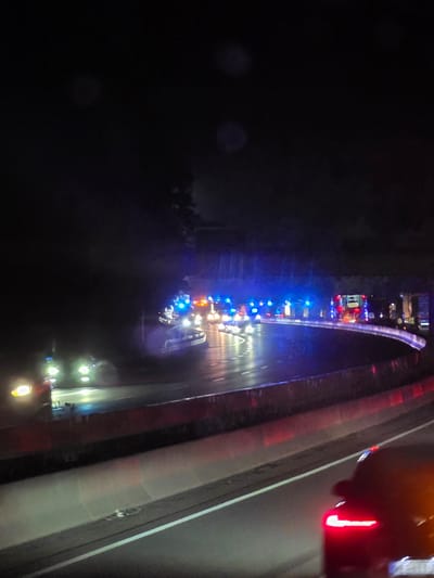 The image depicts a nighttime scene on a highway in or near Seligenstadt, Germany. A line of emergency vehicles, including what appear to be police cars with flashing blue lights and a fire truck with flashing red and blue lights, are stopped on the curving highway. Their lights illuminate the wet roadway and surrounding trees. A few passenger vehicles are also visible, including one in the foreground with its brake lights illuminated. The presence of emergency vehicles suggests an incident or traffic stop has occurred.