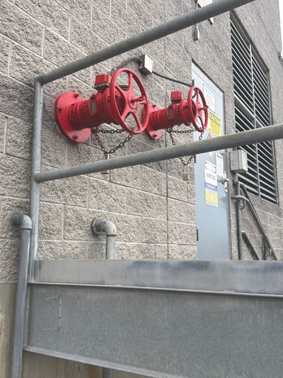 The image depicts a section of an industrial building's exterior wall in Murfreesboro, United States of America. The wall is constructed of light gray, textured brickwork. Two large, red industrial valves with prominent handwheels are mounted to the wall, each secured with chains and padlocks. One valve is labeled with "2000" and what appears to be a manufacturer's name partially obscured. A horizontal galvanized metal pipe runs above the valves, supported by vertical pipes and brackets. To the right of the valves, a gray metal door is visible, adorned with several safety and informational signs. One sign prominently displays "DANGER AMMONIA" and "WARNING." Above the door, a large window or vent with horizontal silver louvers is present. In the foreground, a section of a grey metal structure, possibly stairs or a platform, occupies the lower portion of the frame. The environment appears to be an outdoor industrial setting, with no people visible in the frame.