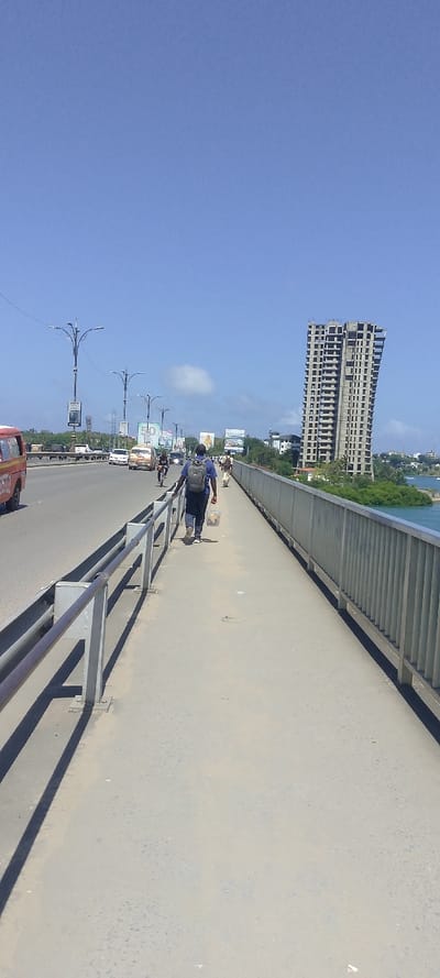 The image captures an elevated roadway and pedestrian bridge in Junda ward, Kenya, under a clear blue sky with minimal scattered clouds. The scene is viewed from a ground-level perspective, looking along the bridge's length.
On the left, a multi-lane asphalt road carries vehicular traffic, including a red van in the foreground, a light-colored public service vehicle (matatu), and other automobiles further down. A cyclist is also visible on the roadway. The road is lined with multiple grey lamp posts featuring multi-arm designs and attached banners or advertisements.
Parallel to the road on the right is a concrete pedestrian sidewalk, separated by a grey metal railing. A person, dressed in a blue shirt, dark trousers, and a grey backpack, walks away from the camera on this sidewalk, carrying a clear plastic bag in their right hand. Further ahead on the same sidewalk, another individual in light-colored clothing is also walking. The outer edge of the pedestrian path is bordered by another grey metal railing.
Beyond the outer railing, a body of blue water, consistent with an estuary or channel, is visible. Green vegetation, likely mangroves, lines the immediate banks of the water. In the background, a prominent unfinished multi-story concrete building with numerous visible structural openings stands tall, indicating ongoing construction. Distant structures and land are also discernible. The environment is brightly lit, casting pronounced shadows from the railings onto the sidewalk.