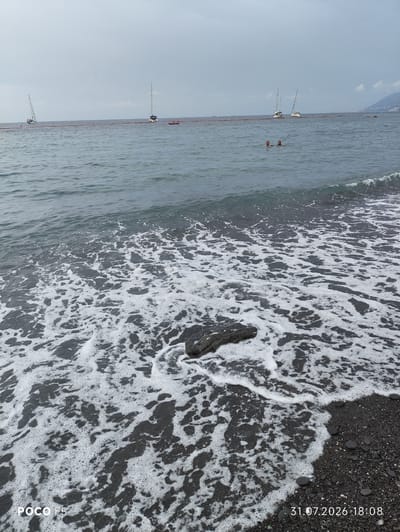 A wide shot captures a choppy grey sea with white foam lapping at a dark pebble beach. Several sailboats are visible on the horizon, anchored in a calm area marked by a red barrier floating on the water. Two individuals are swimming in the sea, their heads and shoulders above the water. The sky is overcast, and a misty mountain range is visible in the far distance on the right. The bottom left corner of the image displays "POCO F5" and the bottom right corner has the date "31.07.2026" and time "18:08". The scene suggests an outdoor recreational area, possibly a beach along the Ligurian coast of Italy given the presence of sailboats and the mountainous backdrop.