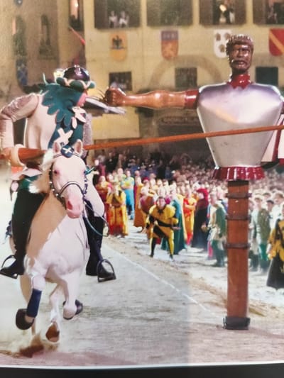The image captures a scene from the Giostra del Saracino in Arezzo, Italy, a historical jousting tournament. In the foreground, a person dressed in historical costume, including a helmet with a green plume and a white and green tunic, rides a white horse. The rider is holding a lance, aimed towards a large, silver, mannequin-like figure with a bronze head and mustache. This figure represents a Saracen and is positioned on a wooden pole. The background is filled with a crowd of spectators, also in colorful, historical attire, observing the event. The architecture in the background suggests a medieval or Renaissance setting, consistent with the historical context of the joust. The ground appears to be a dusty or sandy arena.