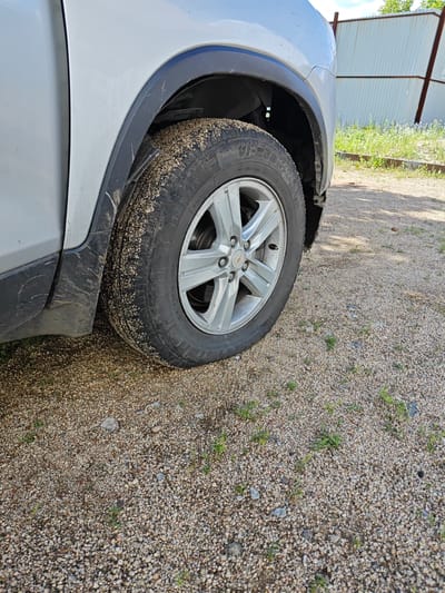 A silver car is parked on a gravel road. The front right tire is visible, covered in dirt and small stones. The wheel is silver with five spokes and a Chevrolet logo in the center. The wheel well is also dirty. In the background, there is a white corrugated metal fence and green grass. The scene appears to be in a rural or industrial area. The location is identified as Oqjetpes, Kazakhstan.