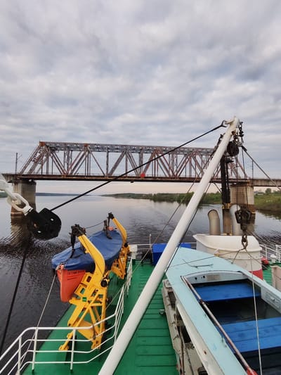 View from a vessel moving on a body of water with a railway bridge in the background. The bridge is a metal truss structure with brown and red coloring, supported by concrete piers. On the vessel's deck, there is an orange lifeboat covered with a blue tarp and a blue rowboat. The deck is green. A white pole and ropes are part of the vessel's equipment. The water is calm with some ripples. The sky is overcast with gray clouds. The location is Tarkanovo, Russia.