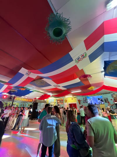 The image displays an indoor scene within what appears to be a lively bar or club, teeming with people gathered for an event. The perspective is oriented as if looking across a room from the left towards a stage or performance area on the right.
The ceiling is heavily decorated with numerous flags, prominently featuring the national flag of the Dominican Republic, distinguishable by its red, white, and blue quadrants and central coat of arms. Interspersed among the flags are various suspended decorative elements, including green, pink, and yellow ornaments.
A significant crowd of people, primarily adults, occupies the right side of the frame. Many are seen from behind or from their profiles, standing and observing an area that appears to be a stage, illuminated by colored lights in shades of pink, blue, and yellow projecting onto the floor. Attendees are dressed casually, suggesting a relaxed, social atmosphere.
On the far right, a clear sign identifies the venue as "COCO BONGO BAR." The walls are adorned with various decor, including large screens displaying images, one notably featuring a white castle or building. The overall lighting is dim and atmospheric, characteristic of an evening or nighttime event. The scene suggests a social gathering at a themed bar or club, likely anticipating or watching a performance.