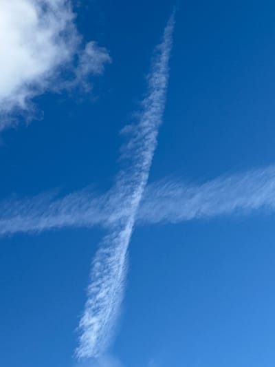 The image displays a clear blue sky with two contrails crossing each other, forming an 'X' shape. One contrail runs diagonally from the bottom left to the top right of the frame, while the other runs vertically from the top to the bottom. The contrails are wispy and appear to be dissipating, suggesting they were formed by aircraft. In the bottom left corner, a small portion of a fluffy white cloud is visible. The scene is set outdoors, with no specific location cues other than the provided context of Vsevolodovo, Russia. The time of day appears to be daytime, given the bright blue sky. There is no activity or event taking place, beyond the natural formation and dissipation of contrails. No people, objects, or text are visible in the image.