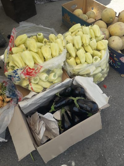 This overhead shot captures a vibrant display of fresh produce at what appears to be an outdoor market or roadside stall in Ghafurov, Tajikistan. The items are laid out on a dark gray asphalt or concrete surface under bright daylight, suggested by the clear illumination and visible shadows.

The main subjects are various fruits and vegetables. To the left, a wooden crate holds several light yellow or pale green melons, with more piled loosely beside it. These melons are round to oval in shape.

Dominating the center of the image are large quantities of pale yellow bell peppers, stored in clear plastic bags. There are at least two substantial bags filled with these peppers, one appearing fuller than the other.

To the right, a cardboard box contains numerous dark purple, almost black, eggplants. They are arranged within the box, with some white crumpled paper serving as packing material. Below this box, in the bottom right corner, another partially visible clear plastic bag holds light brown root vegetables, possibly potatoes or onions, some of which show green sprouts.

The setting is simple, with the produce displayed directly on the ground in various containers typical of market settings. No people are visible, so there are no discernible emotions.

Visible text includes partial brand names or labels on the packaging. On the blue side of a melon crate, parts of words like "ADOR" and "VERA" or "VEGA" can be seen. On the plastic bags holding the bell peppers, some red text is visible, including fragments like "AIAA," "POK," and "KHIVAK" or "SHIVAKI".