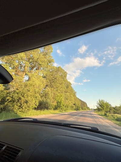 The image, taken from inside a vehicle, depicts a rural road leading into a forest. The road surface is unpaved and appears to be dirt or gravel. Lush green trees line both sides of the road, with sunlight filtering through their leaves, casting dappled shadows. The sky is a clear blue with scattered white clouds. In the distance, a power line pylon is visible. The interior of the car, including the dashboard and windshield wipers, is visible in the foreground. The scene suggests a drive through a natural landscape on a sunny day. The location is identified as Ihrovytsia, Ukraine.